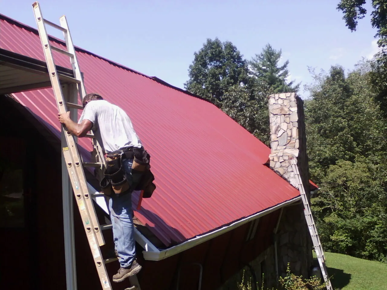 Roofer on a ladder beside a red metal roof and stone chimney — Soffit & Fascia Repair by Roofing Brothers in Lambertville