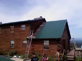 Green metal roof going on a two story log home with ladders set in Lambertville
